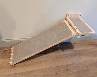 May include: A wall-mounted cat furniture set consisting of a rectangular wooden platform and a long, angled wooden ramp. Both pieces feature light-colored wood frames and are inlaid with textured gray fabric panels, connected by thick white rope.