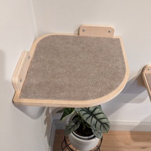 May include: A light brown, rounded, wooden cat shelf with a beige carpet surface. The shelf is mounted on a white wall and is designed for a corner.