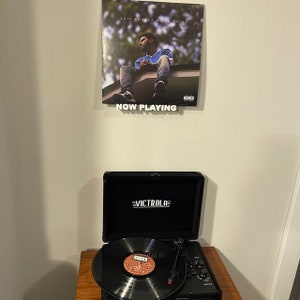 May include: A black record player with a record on the turntable. The record is a vinyl record with a black and white image of a person on the cover. The record player is on a wooden surface. The record player has a black case with the word "VICTROLA" printed in white on the front. The record player is playing a record with the album cover "2014 Forest Hills Drive" by J. Cole.