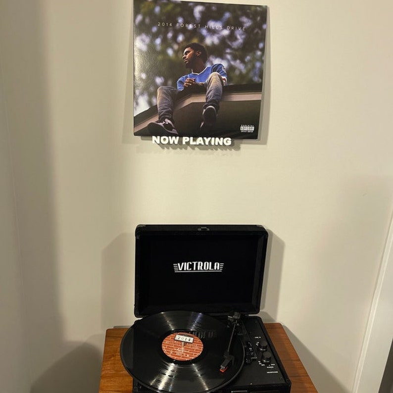 May include: A black record player with a record on the turntable. The record is a vinyl record with a black and white image of a person on the cover. The record player is on a wooden surface. The record player has a black case with the word "VICTROLA" printed in white on the front. The record player is playing a record with the album cover "2014 Forest Hills Drive" by J. Cole.