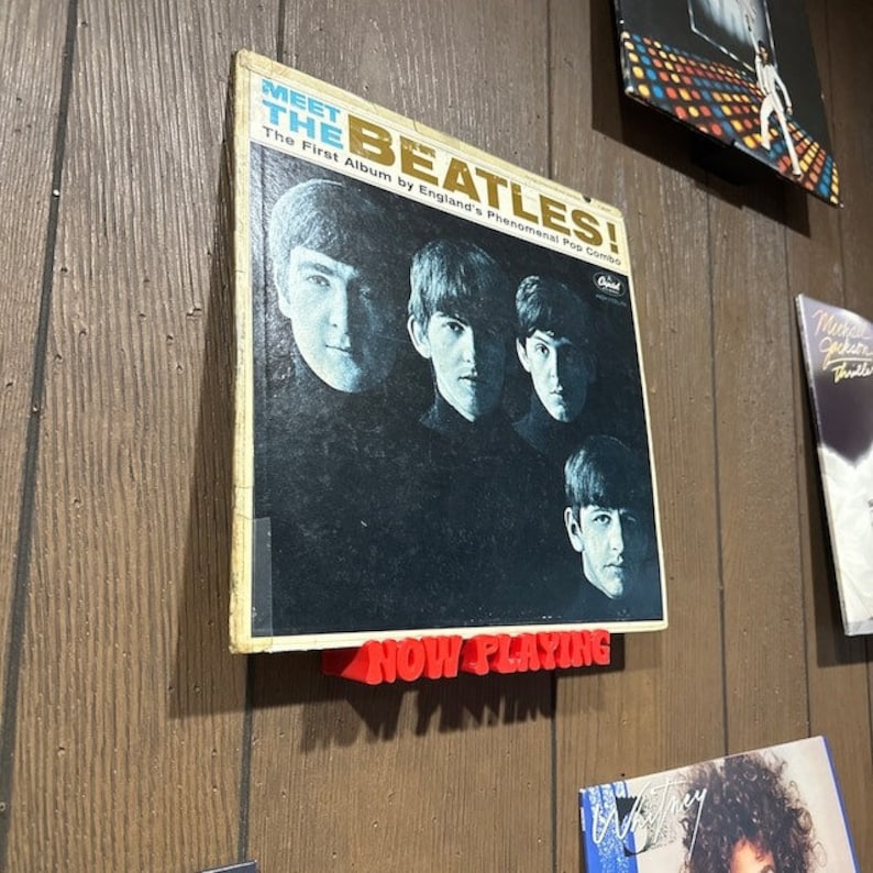 May include: A vintage vinyl record album cover featuring a black and white photo of the Beatles with the text "Meet The Beatles! The First Album by England's Phenomenal Pop Combo" and a red "Now Playing" sign.