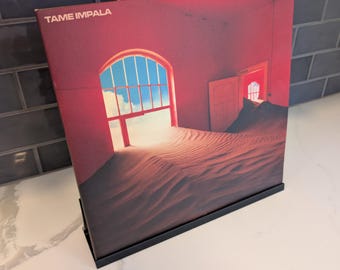 May include: A square vinyl record jacket for Tame Impala's The Slow Rush. The cover art features a red-toned room filled with sand, an open arched window showing a blue sky, and an open door. The text TAME IMPALA and THE SLOW RUSH is visible.
