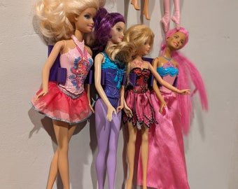 May include: Five Barbie dolls with different outfits and hairstyles are hanging on a white wall. The dolls are wearing pink, red, purple, and blue dresses. They have long blonde, purple, and pink hair.