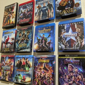 May include: A wall shelf with a collection of Marvel movie Blu-ray discs. The discs are arranged in rows and include titles such as Captain America, Avengers, Spider-Man, Guardians of the Galaxy, Black Panther, Doctor Strange, and Civil War.