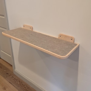 May include: A wall-mounted cat shelf with a light gray felt surface and natural wood trim. The shelf is rectangular with rounded corners and is attached to the wall with two wooden brackets.