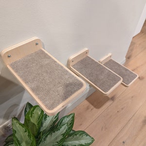 May include: Three wooden cat shelves mounted on a wall. Each shelf has a light-colored wood frame and a rectangular, carpeted surface. The shelves are staggered in size and position, creating a climbing structure for cats. The shelves are attached to the wall with screws.