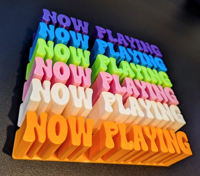 May include: A stack of colorful blocks spelling out "NOW PLAYING" in a rainbow of colors, including purple, blue, green, pink, white, and orange.