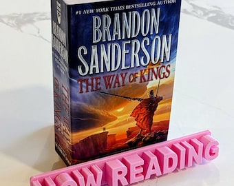 May include: A pink bookend with the words "NOW READING" in raised letters. The bookend is in front of a paperback book titled "The Way of Kings" by Brandon Sanderson.