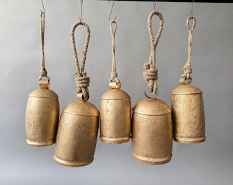 May include: Five gold-colored metal bells hanging from a metal bar with natural rope hangers. The bells have a rustic, textured finish and vary slightly in size and shape. The rope is knotted at the top.