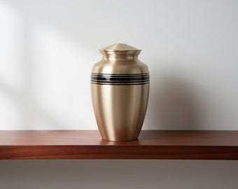 May include: A gold-colored urn with a rounded shape and a lid. The urn features two black bands around the middle. It is displayed on a dark brown wooden shelf against a white wall.