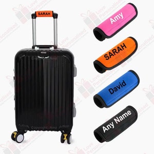 May include: A black travel suitcase with a pull-out handle, displaying the name "SARAH". The image also shows handle wraps in pink, orange, blue, and black, each with a name: "Amy", "SARAH", "David", and "Any Name".