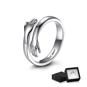 May include: A silver ring featuring a design of two hands in an embrace. The ring is engraved with the words "Text Here". The ring is presented in a black gift box with a black ribbon.