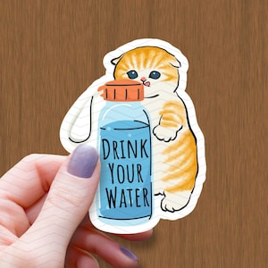 Könnte beinhalten: Ein Aufkleber mit einem Cartoon-Katzenbild eines orangefarbenen Tabbys, der eine blaue Wasserflasche mit dem Text "Drink Your Water" hält.