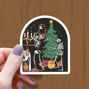 Op de afbeelding: Twee skeletten versieren een kerstboom met ornamenten en een ster bovenop. De boom is groen met rode en gouden ornamenten. De skeletten dragen een kerstmanmuts en een schedel. De scène speelt zich af in een donkere kamer met een open haard en kaarsen.
