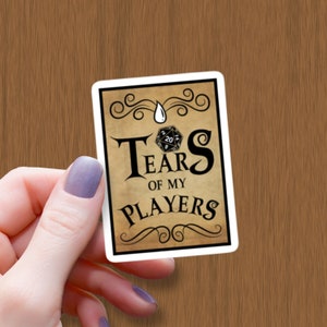 May include: A white sticker with a brown background and black text that reads "Tears of my Players" with a black and white 20-sided die in the centre.