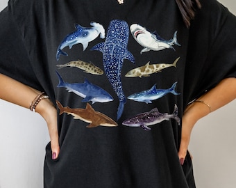 May include: Black t-shirt featuring a graphic print of various shark species, including a whale shark, hammerhead shark, and a whitetip reef shark.