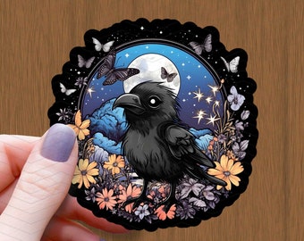 May include: A black raven with large eyes sits in a circle of flowers and stars against a blue night sky with a full moon. The raven is surrounded by a variety of flowers in shades of yellow, pink, and purple. The moon is surrounded by stars and a few butterflies.