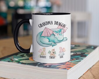 May include: White ceramic mug with a black rim and handle. The mug features a watercolor illustration of a sleeping blue dragon with pink wings. The text "Grandma Dragon" is printed above the dragon. Below the dragon are three smaller dragons with the names "Bobby", "Trish", and "Sue" printed underneath.