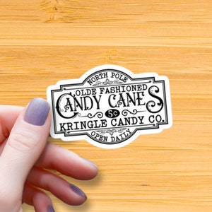 May include: A white sticker with black text that reads "North Pole Olde Fashioned Candy Canes 5¢ Kringle Candy Co. Open Daily". The sticker has a vintage design with ornate lettering and a decorative border.