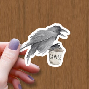 May include: A black and grey crow with a white cup that says "CAWFFEE" in black lettering. The crow is holding the cup with its feet.