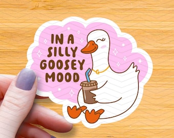 May include: A sticker featuring a cartoon goose with a coffee cup and the phrase "IN A SILLY GOOSEY MOOD." The goose is white with an orange beak and feet, wearing a gold necklace. The background is pink with white stars.