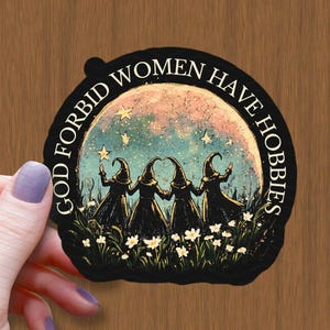 Puede incluir: Una pegatina negra con el texto "GOD FORBID WOMEN HAVE HOBBIES" que rodea una ilustración de cuatro brujas con túnicas y sombreros negros, de pie en un campo de flores blancas, con una luna y estrellas en el fondo.