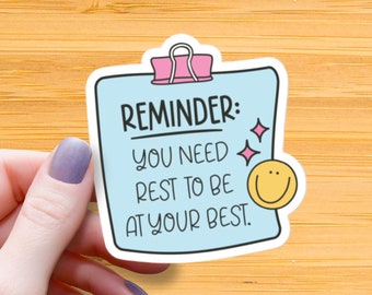 Può includere: Un adesivo blu e bianco con un promemoria per riposare per essere al meglio. L'adesivo ha una graffetta rosa e un'emoticon sorridente.