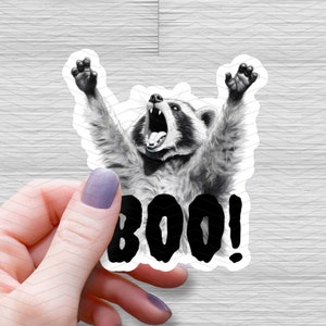 May include: A black and white sticker of a raccoon with its mouth open, as if screaming, with the word "BOO!" in black letters below it.