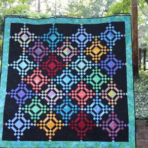Puede incluir: Una colcha colorida con un patrón geométrico de cuadrados y diamantes. La colcha está hecha de varios tonos de azul, verde, rojo, naranja y morado. La colcha está colgada en un tendedero en un patio trasero.
