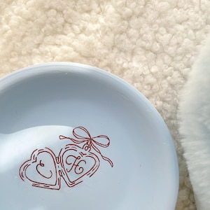 May include: A light blue ceramic plate with a red heart-shaped design. The design features two connected hearts with the letter "H" inside one heart and a bow at the top. The plate is on a textured, off-white surface.