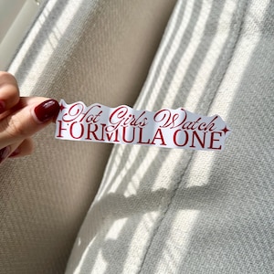 Op de afbeelding: Een witte sticker met een geschulpte rand, met de tekst "Hot Girls Watch FORMULA ONE" in rood script. De sticker wordt vastgehouden door een hand met rode nagellak, tegen een neutrale achtergrond.