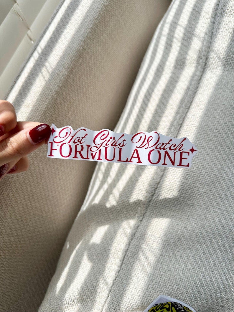 Op de afbeelding: Een witte sticker met een geschulpte rand, met de tekst "Hot Girls Watch FORMULA ONE" in rood script. De sticker wordt vastgehouden door een hand met rode nagellak, tegen een neutrale achtergrond.