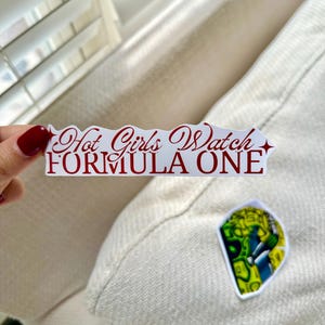 Op de afbeelding: Een witte sticker met de tekst "Hot Girls Watch FORMULA ONE" in rood script. Een kleinere sticker met een kleurrijk ontwerp is ook zichtbaar.