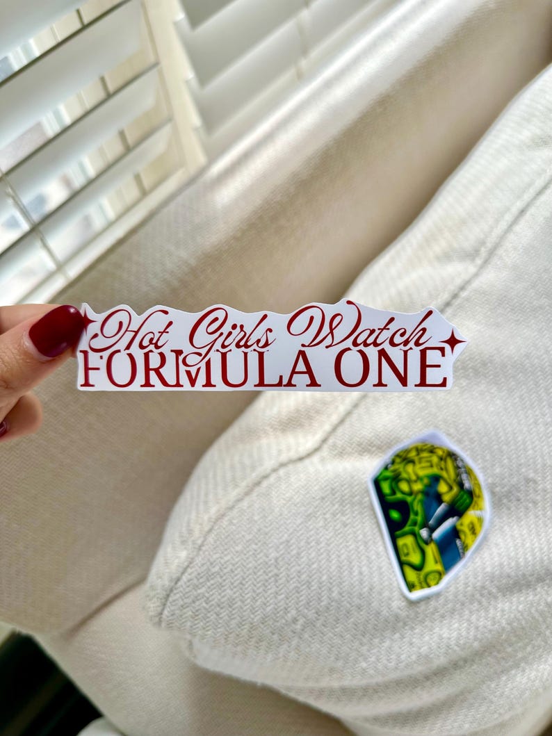 Op de afbeelding: Een witte sticker met de tekst "Hot Girls Watch FORMULA ONE" in rood script. Een kleinere sticker met een kleurrijk ontwerp is ook zichtbaar.