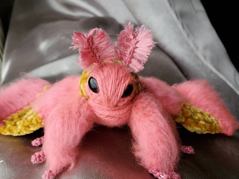 May include: A pink and yellow crocheted moth plush toy with fuzzy pink fur and yellow velvet wings.