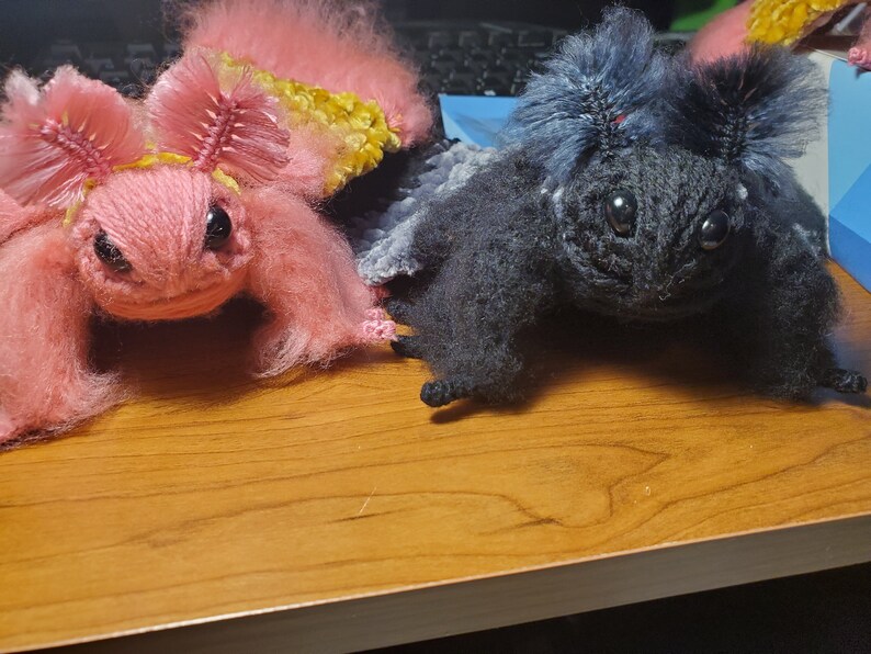 May include: Two crocheted plush toys, one pink with yellow accents and one black, with large eyes and fuzzy textures.