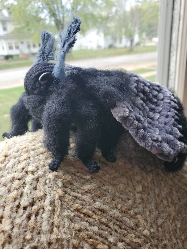 May include: A black and gray plush moth with fuzzy wings. The moth is sitting on a brown knitted surface.