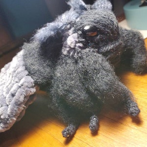 May include: A black and gray crocheted plush toy with a long, furry tail and large, black eyes. The toy has a textured, fuzzy surface and is posed with its wings spread out.
