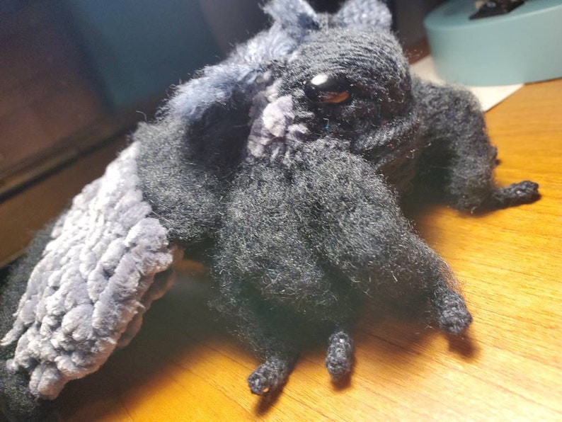 May include: A black and gray crocheted plush toy with a long, furry tail and large, black eyes. The toy has a textured, fuzzy surface and is posed with its wings spread out.