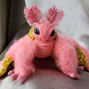 May include: A pink and yellow crocheted plush toy resembling a moth with fuzzy wings and large eyes.