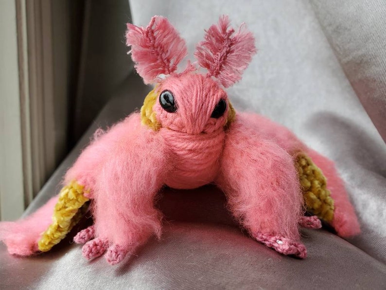 May include: A pink and yellow crocheted plush toy resembling a moth with fuzzy wings and large eyes.