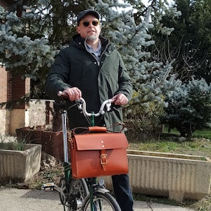 Bolso de cuero para bicicletas Brompton grande (para tablets,Macbook pro 13"...) colocado en la bicicleta Brompton con modelo masculino en un jardín.Modelo de cuero marrón intermedio.