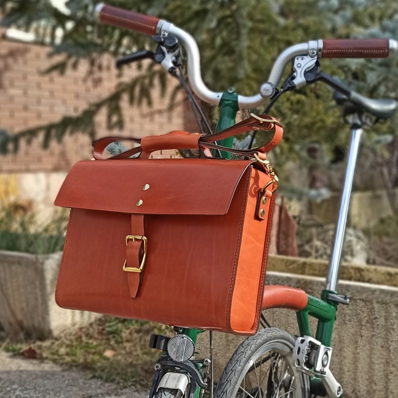 Bolso de cuero para bicicletas Brompton grande (para tablets,Macbook pro 13"...) colocado en la bicicleta Brompton en un jardín.Modelo de cuero marrón intermedio. Vista latero-frontal. Se aprecia además la bandolera recogida en el asa.