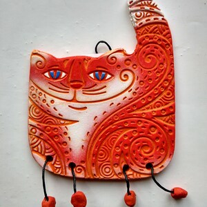 May include: Orange and white ceramic cat figurine with intricate patterns and blue eyes. The cat has a raised tail and is hanging from black cords with orange beads.