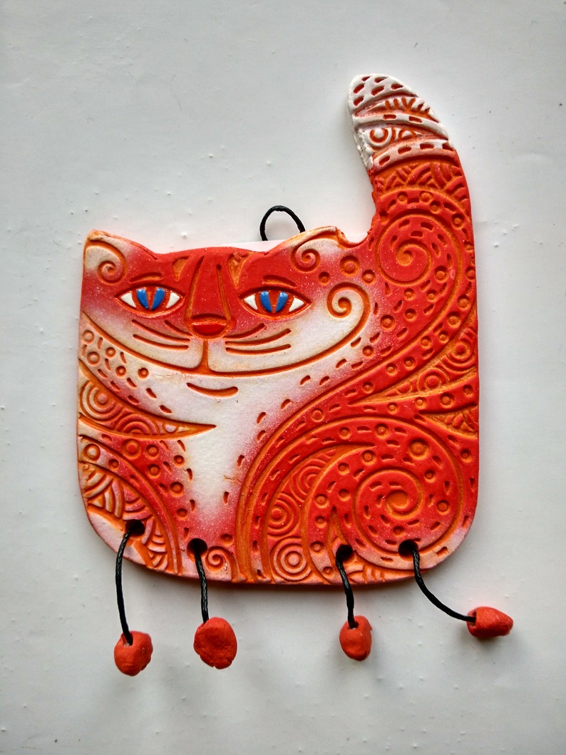 May include: Orange and white ceramic cat figurine with intricate patterns and blue eyes. The cat has a raised tail and is hanging from black cords with orange beads.