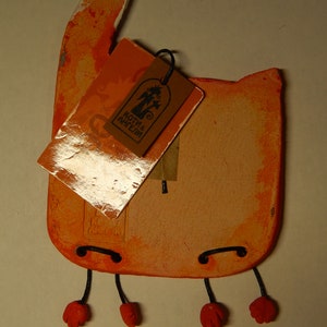 May include: Orange ceramic cat figurine with black string legs and red feet. The cat is holding a tag that says "Коты и Ангелы".