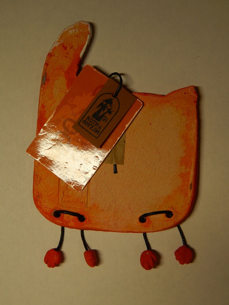 May include: Orange ceramic cat figurine with black string legs and red feet. The cat is holding a tag that says "Коты и Ангелы".
