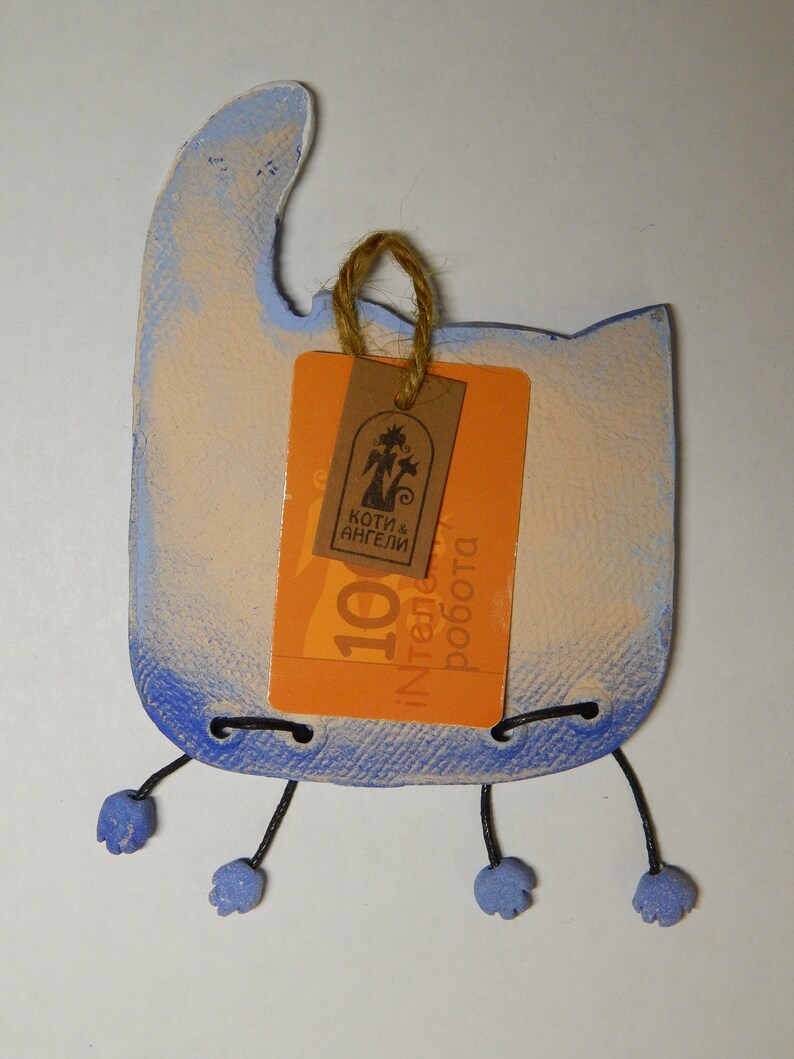 May include: A blue and white ceramic cat figurine with a tag that reads "Koti & Angeli" and "100% ręczna robota". The cat has black string legs and blue feet.