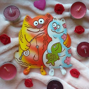 Puede incluir: Una caprichosa figura de gato de cerámica en naranja y azul, sosteniendo una caña de pescar y un pez verde. Rodeada de rosas rojas, un corazón rosa y velas de té sobre una superficie rosa. Artículo decorativo.