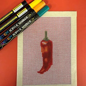 May include: A red chili pepper needlepoint design on a white grid canvas, with a beige border. Four acrylic markers in red, yellow, orange, and teal are arranged diagonally. The markers have black barrels and the text "ACRYLIC MARKER".